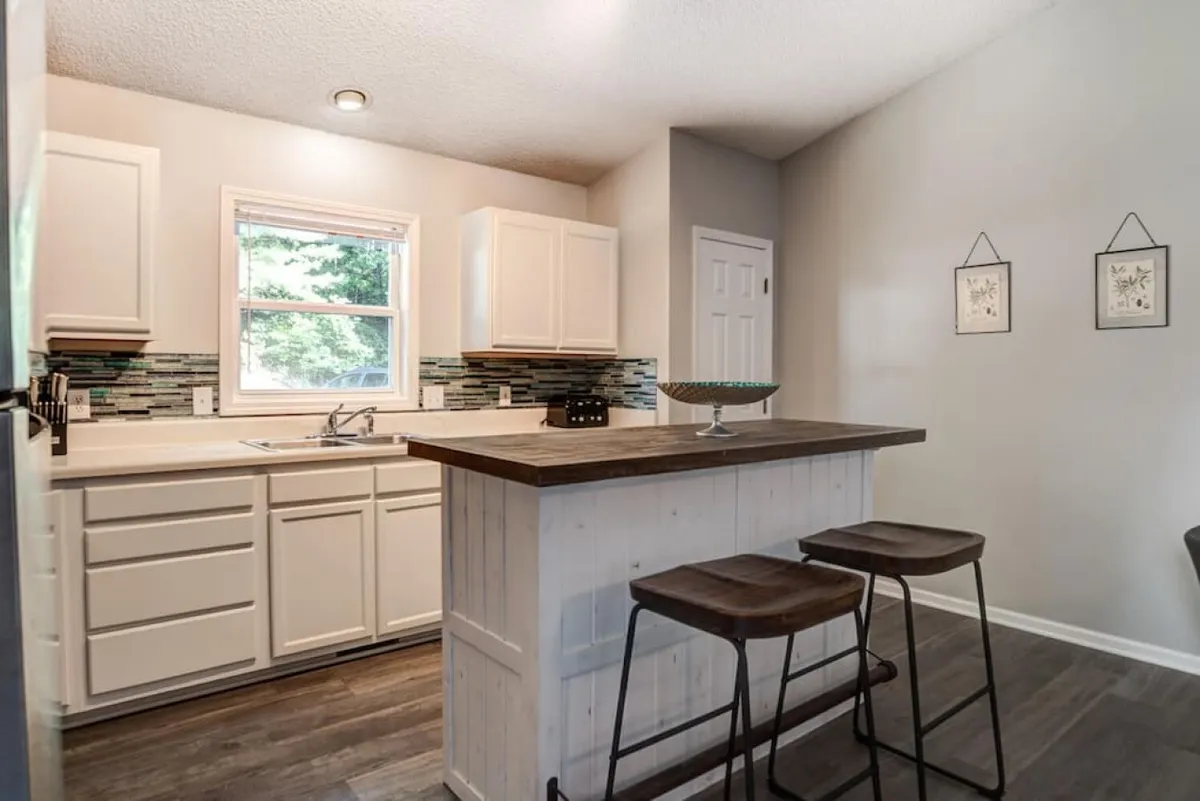 A welcoming kitchen scene with a spacious island and stools, inviting guests to gather and chat while meals are being prepared. A large window lets in natural light, and stylish backsplash tiles add a touch of modern flair. The setup is perfect for casual brunches or holiday celebrations.