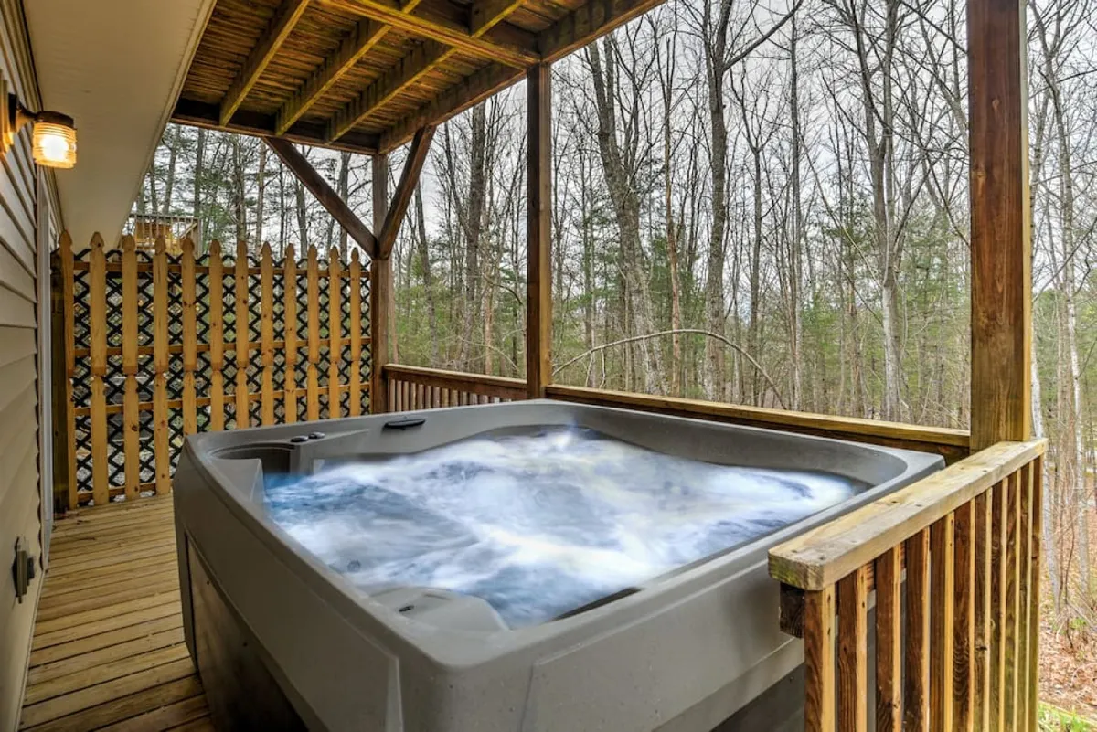 An inviting view of a wooden deck fitted with a hot tub, surrounded by dense, mature trees. The natural privacy and spa feature evoke a sense of retreat and relaxation, with ample room for seating or gathering. The setting is serene, suggesting possibilities for stargazing, morning routines, or unwinding after a day of urban adventure.
