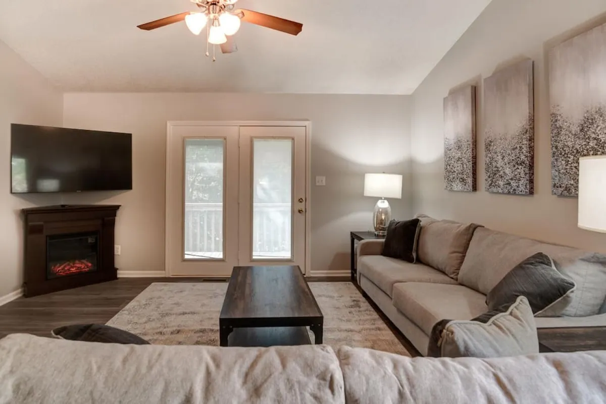 A warm, inviting living room featuring a plush sectional sofa facing a fireplace and television. Sunlight streams through the windows, illuminating tasteful artwork and a coffee table set up for casual gatherings. The layout is open and encourages both relaxation and social interaction.