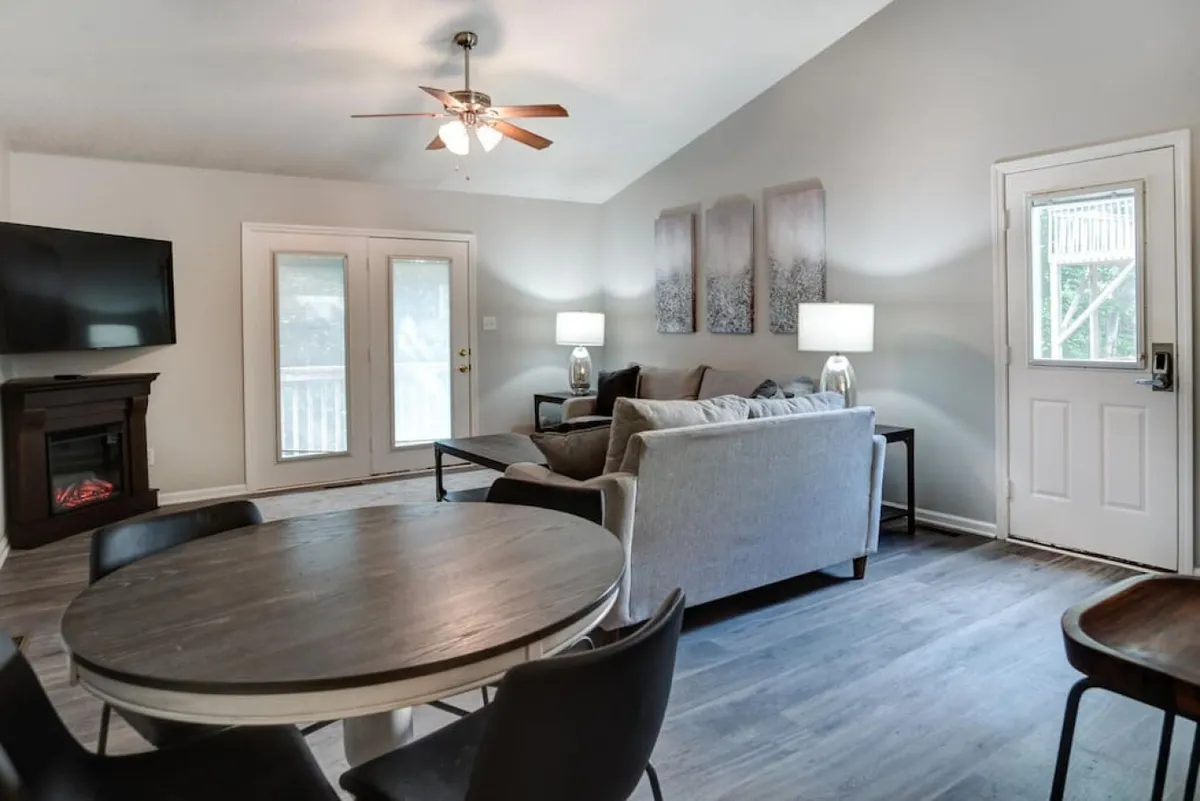 The image showcases a contemporary open-concept living and dining area featuring a stylish dining table set for gathering, a plush sofa set arranged around a modern fireplace, colorful artwork on the wall, and glass doors that open to a sunlit outdoor space.