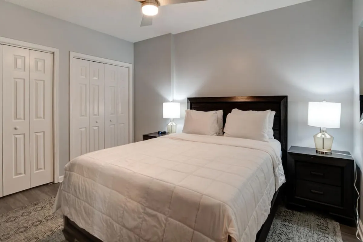 This image showcases a tranquil bedroom with a neatly made bed, nightstands on either side with soft-lit lamps, and visible closet doors. The space is airy and inviting, with soft lighting that enhances the sense of calm and comfort.
