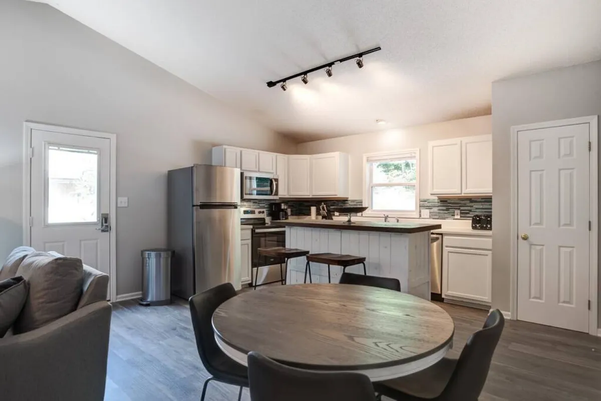 A bright, modern kitchen and dining area highlighted by a central island, circular dining table, and stainless steel appliances. Sunlight bathes the space, making it ideal for shared meals, casual coffee mornings, or baking sessions. The seamless connection to the living area is implied by the open sightlines.