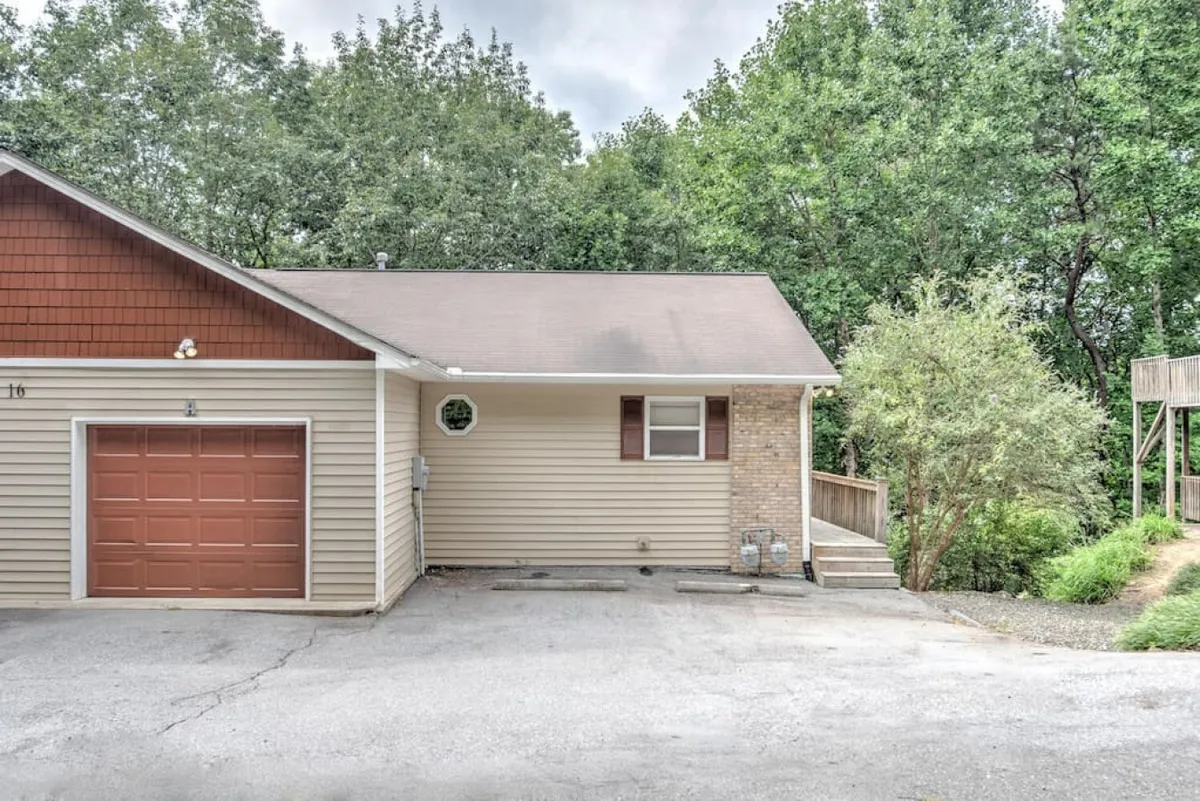 Here, the exterior is shown with a close-up view of the garage door, brick accents, wooden deck, and lush surrounding greenery. The image evokes a welcoming atmosphere and highlights the accessibility of the entry and parking areas.