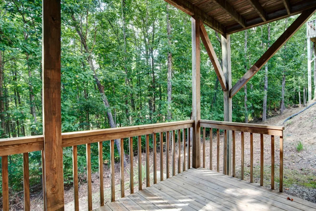 A peaceful exterior scene featuring a private wooden deck bordered by lush green trees. The deck has a sturdy wooden railing and open space, suggesting room for outdoor seating or relaxation. Natural sunlight filters through the trees, creating an inviting spot for leisure or gathering with friends and pets.
